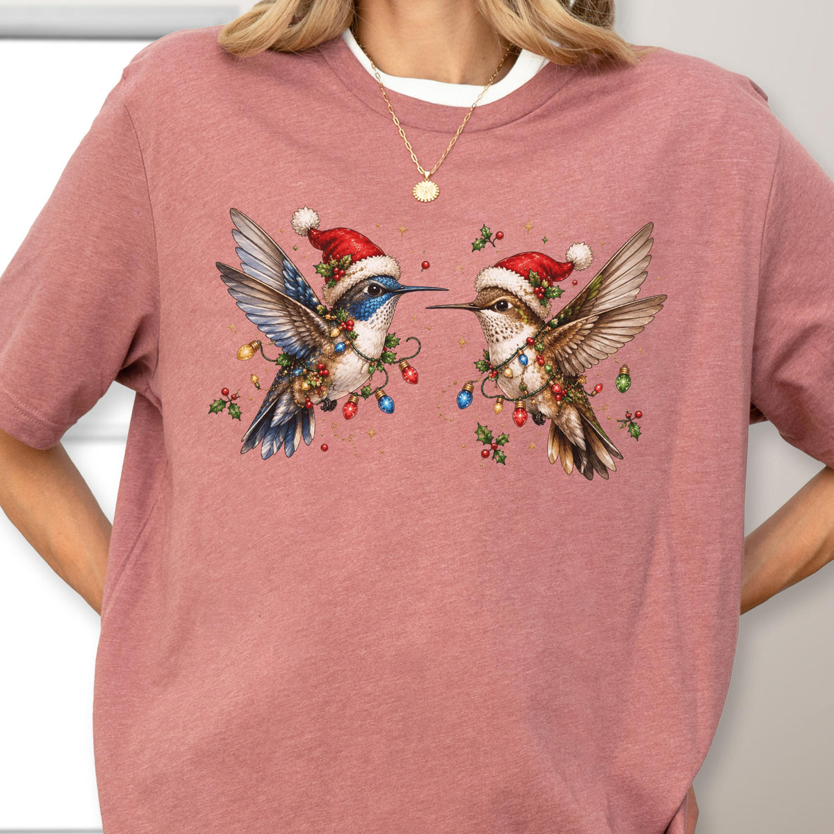 May include: A mauve short-sleeve t-shirt featuring a graphic of two hummingbirds wearing red and white Santa hats. The birds are decorated with colorful Christmas light bulbs and green holly leaves, creating a festive holiday-themed illustration.