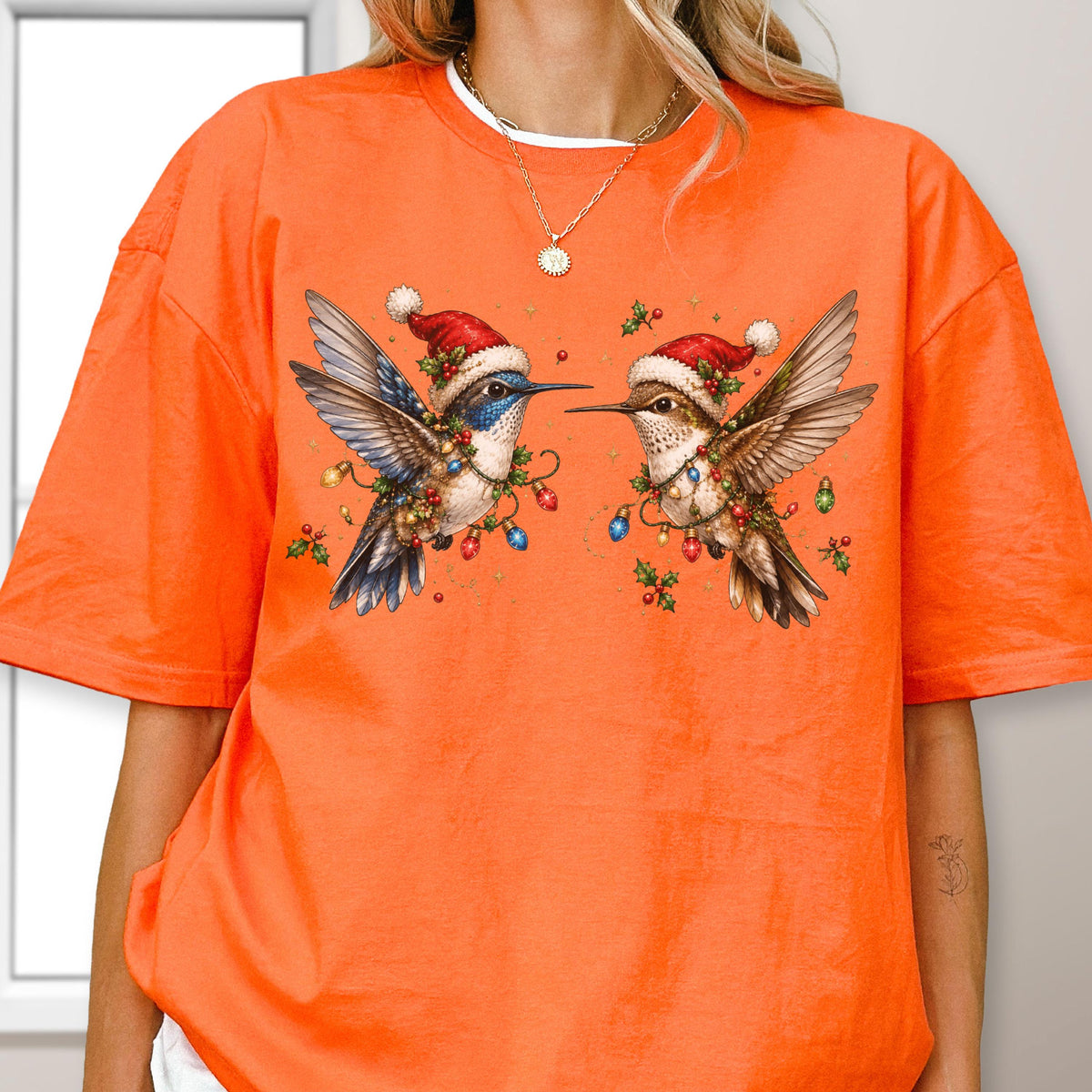 May include: An orange short-sleeved t-shirt featuring a graphic of two hummingbirds wearing red and white Santa hats. The birds are surrounded by colorful Christmas light bulbs and green holly leaves, creating a festive holiday-themed illustration.