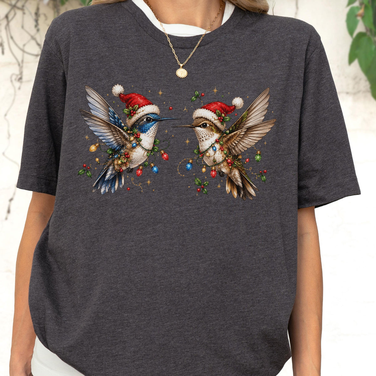 May include: A dark heather gray short-sleeve t-shirt featuring a graphic of two hummingbirds wearing red and white Santa hats. The birds are decorated with colorful Christmas lights and holly berries, with one bird showing blue plumage and the other brown.