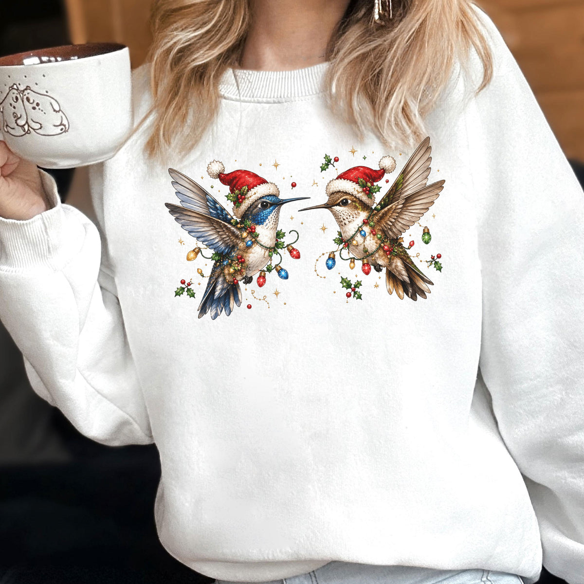 May include: A white crewneck sweatshirt featuring a graphic print of two hummingbirds wearing red Santa hats. The birds are entangled in colorful Christmas light strings and holly berries, with a white mug held nearby showing a line drawing of two bears.
