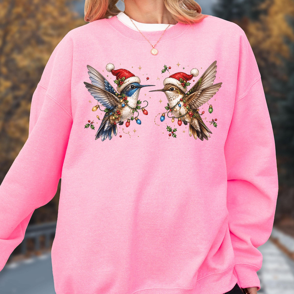 May include: A bright pink long-sleeve sweatshirt featuring a graphic of two hummingbirds wearing red Santa hats. The birds are surrounded by colorful Christmas light bulbs and green holly leaves, creating a festive holiday-themed design.