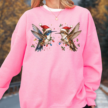 May include: A bright pink long-sleeve sweatshirt featuring a graphic of two hummingbirds wearing red Santa hats. The birds are surrounded by colorful Christmas light bulbs and green holly leaves, creating a festive holiday-themed design.