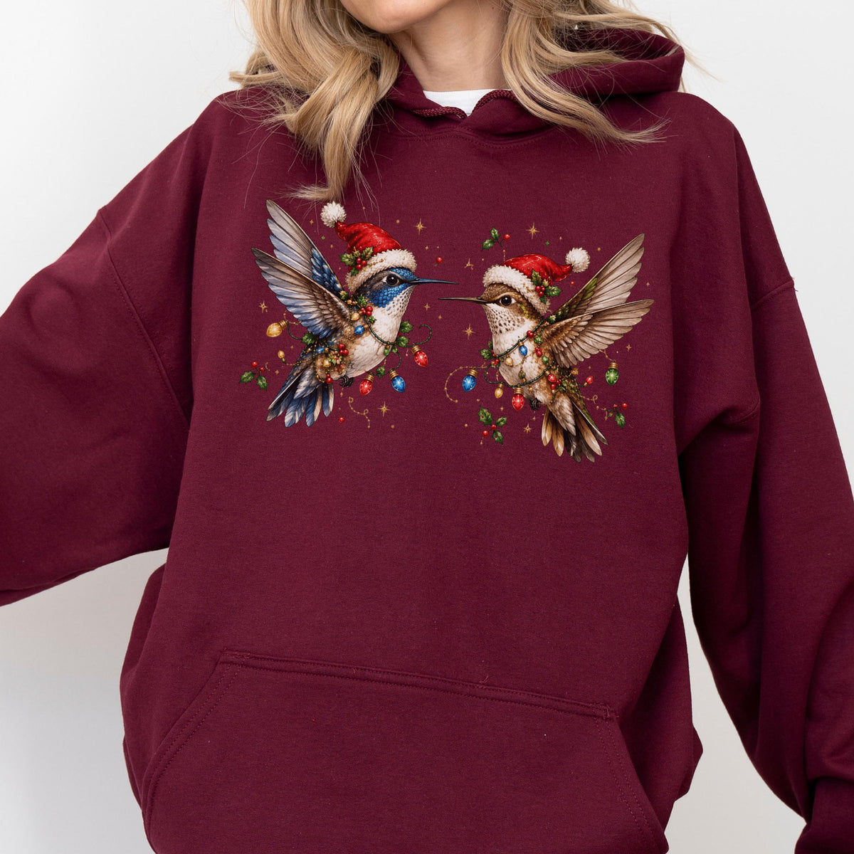 May include: A maroon hooded sweatshirt featuring a graphic print of two hummingbirds wearing red Santa hats. The birds are surrounded by colorful Christmas lights, holly berries, and small gold stars, creating a festive holiday-themed design.