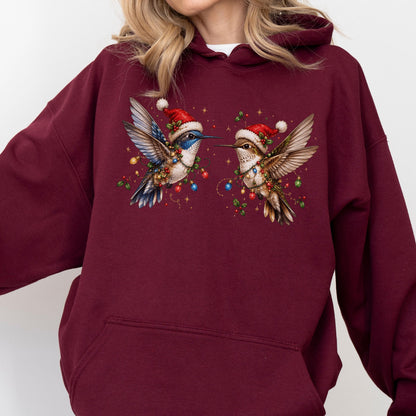 May include: A maroon hooded sweatshirt featuring a graphic print of two hummingbirds wearing red Santa hats. The birds are surrounded by colorful Christmas lights, holly berries, and small gold stars, creating a festive holiday-themed design.