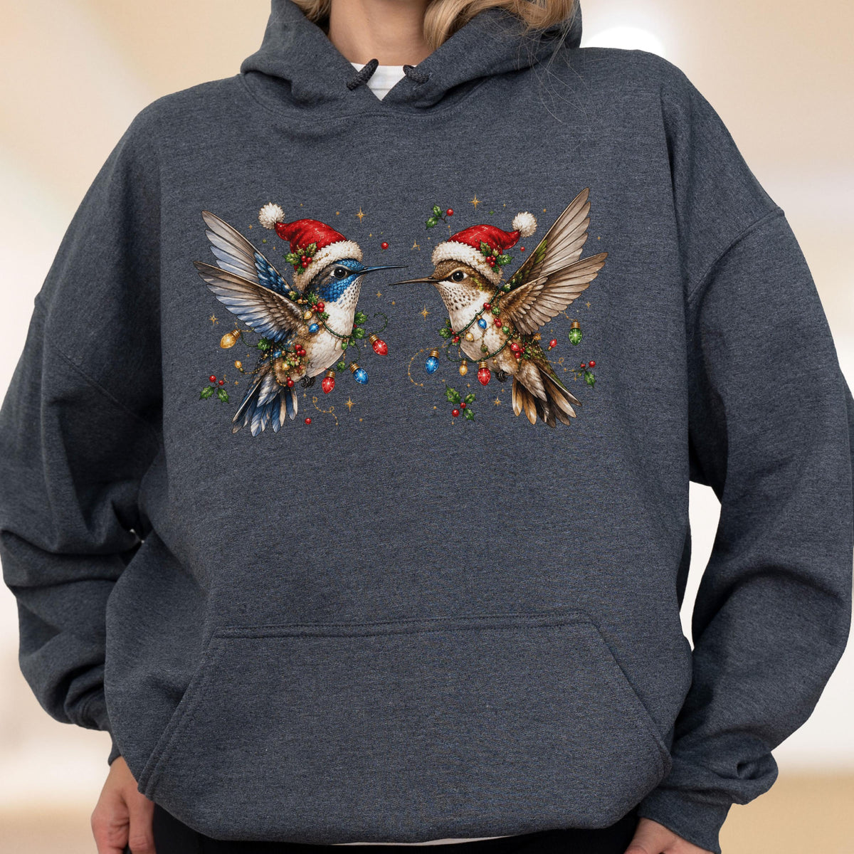 May include: A dark heather gray hooded sweatshirt featuring a graphic print of two hummingbirds wearing red and white Santa hats. The birds are surrounded by colorful Christmas light bulbs, green holly leaves, and red berries on the chest area.