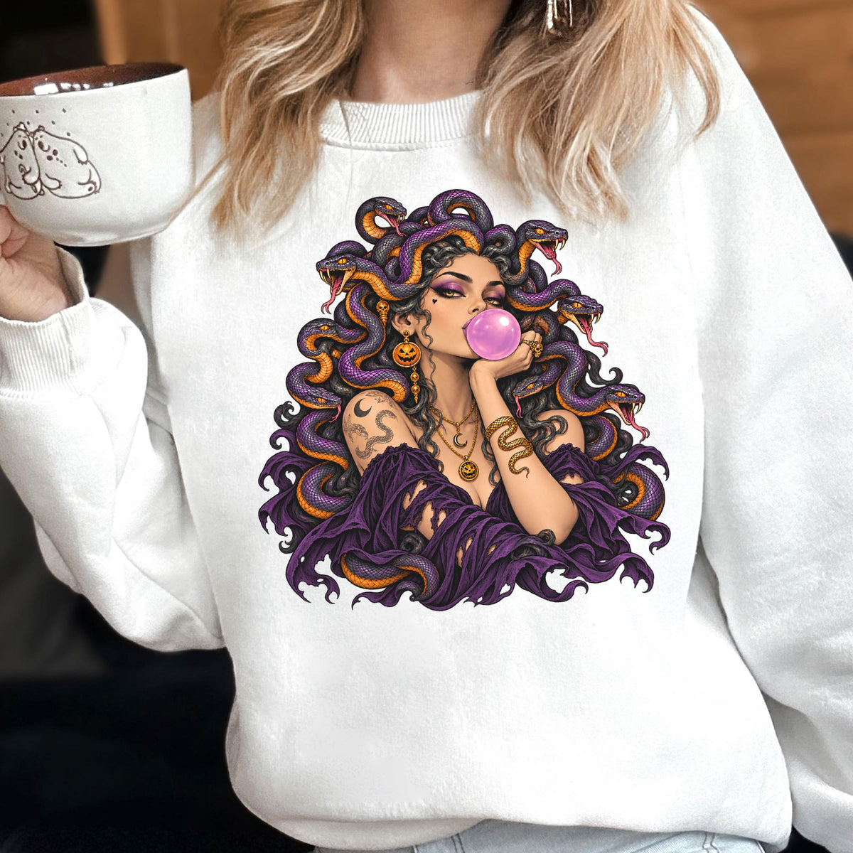 May include: A white crewneck sweatshirt featuring a graphic print of a figure with purple snakes for hair, blowing a pink bubblegum bubble. The figure wears a dark purple off-the-shoulder top, layered gold necklaces, and pumpkin earrings.