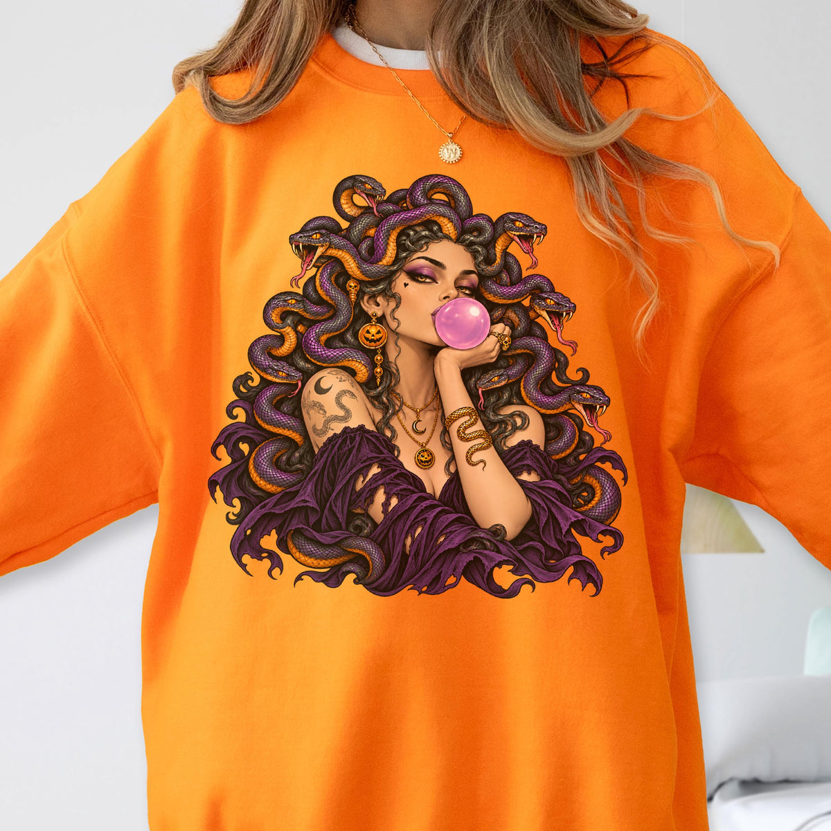 May include: An orange sweatshirt featuring a graphic print of a figure with snakes for hair, wearing a purple garment, and blowing a pink bubble. The figure has gold jewelry, including a necklace and earrings, and a snake-like arm cuff.