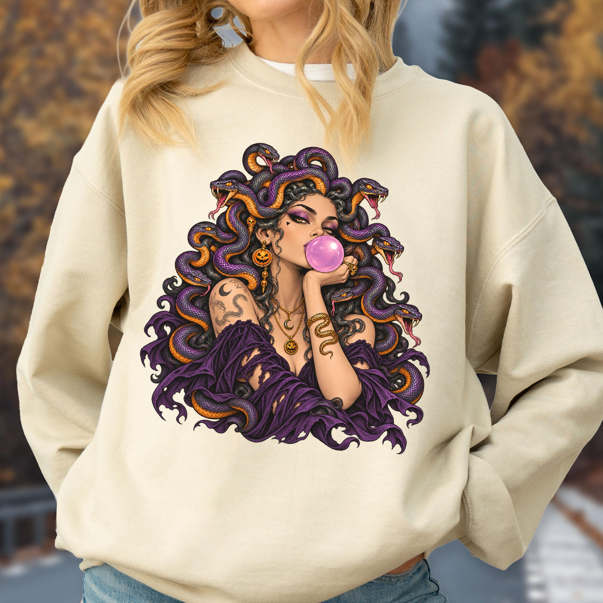 May include: A beige crewneck sweatshirt featuring a graphic print of a figure with purple snakes for hair, wearing a dark purple off-the-shoulder top, gold jewelry, and blowing a pink bubble with gum. The design includes intricate snake details.