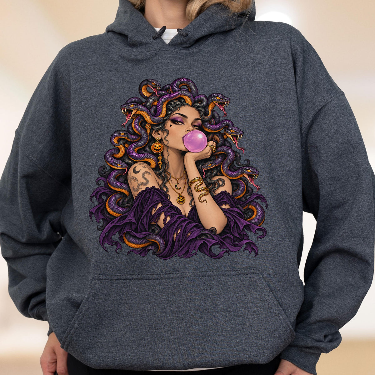 May include: A charcoal gray hooded sweatshirt featuring a graphic print of a figure with snakes for hair, wearing a purple off-the-shoulder top, blowing a pink bubble, and adorned with gold jewelry including a pumpkin pendant and snake arm cuff.