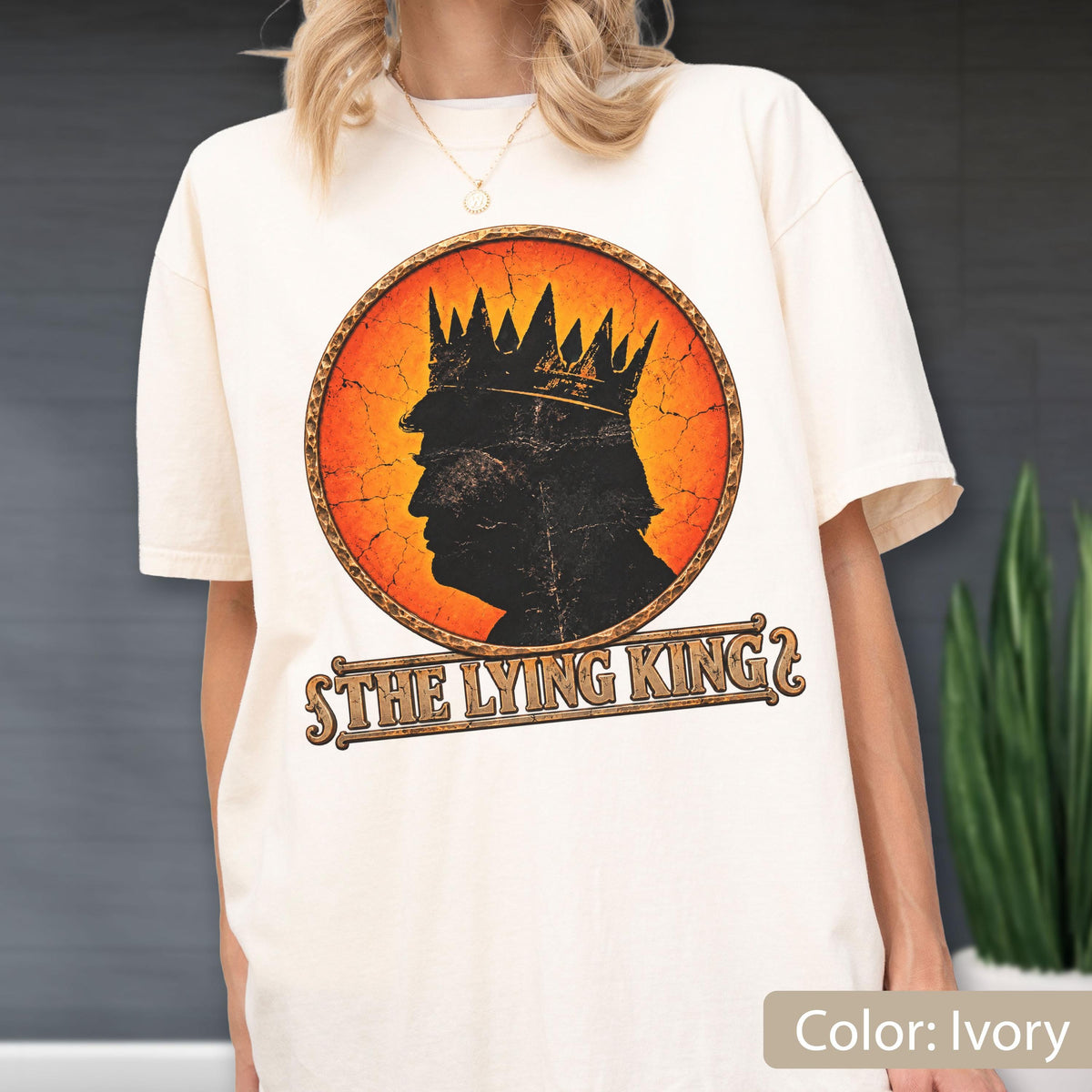 May include: An ivory short-sleeve t-shirt featuring a circular graphic with a distressed orange and yellow background. The graphic depicts a black silhouette of a person wearing a crown. Below the circle, the text reads THE LYING KING.