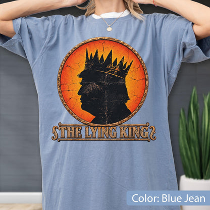 May include: A blue jean colored t-shirt featuring a circular graphic with an orange and red gradient background. The graphic shows a black silhouette of a person wearing a crown, with the text THE LYING KING printed in a decorative font below.