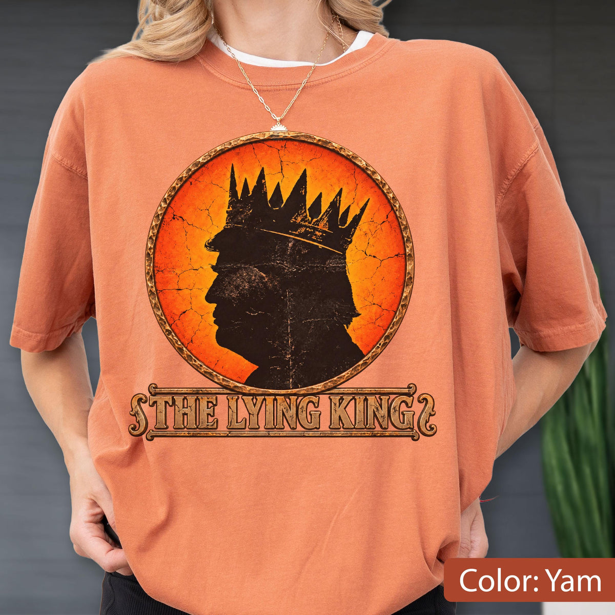 May include: An orange t-shirt featuring a circular graphic with a black silhouette of a person wearing a crown against a cracked orange and yellow background. Below the circle, the text THE LYING KING is printed in a decorative brown font.
