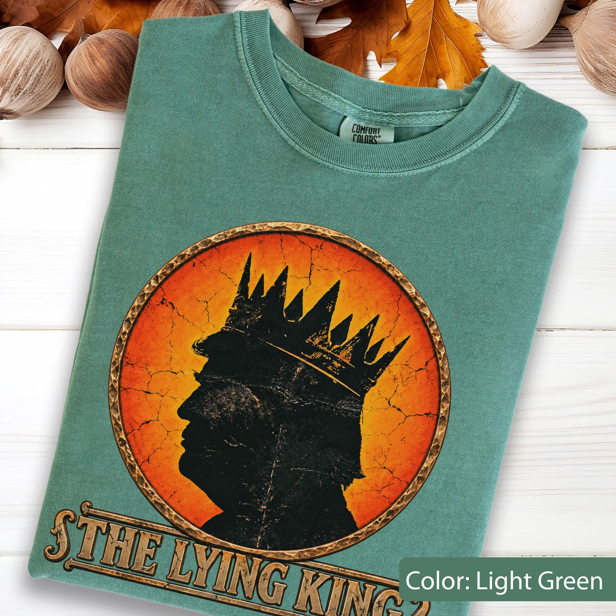 May include: A light green t-shirt featuring a circular graphic with a distressed orange background. The graphic depicts a black silhouette of a person wearing a crown. Below the circle, the text reads "THE LYING KING" in a stylized font.