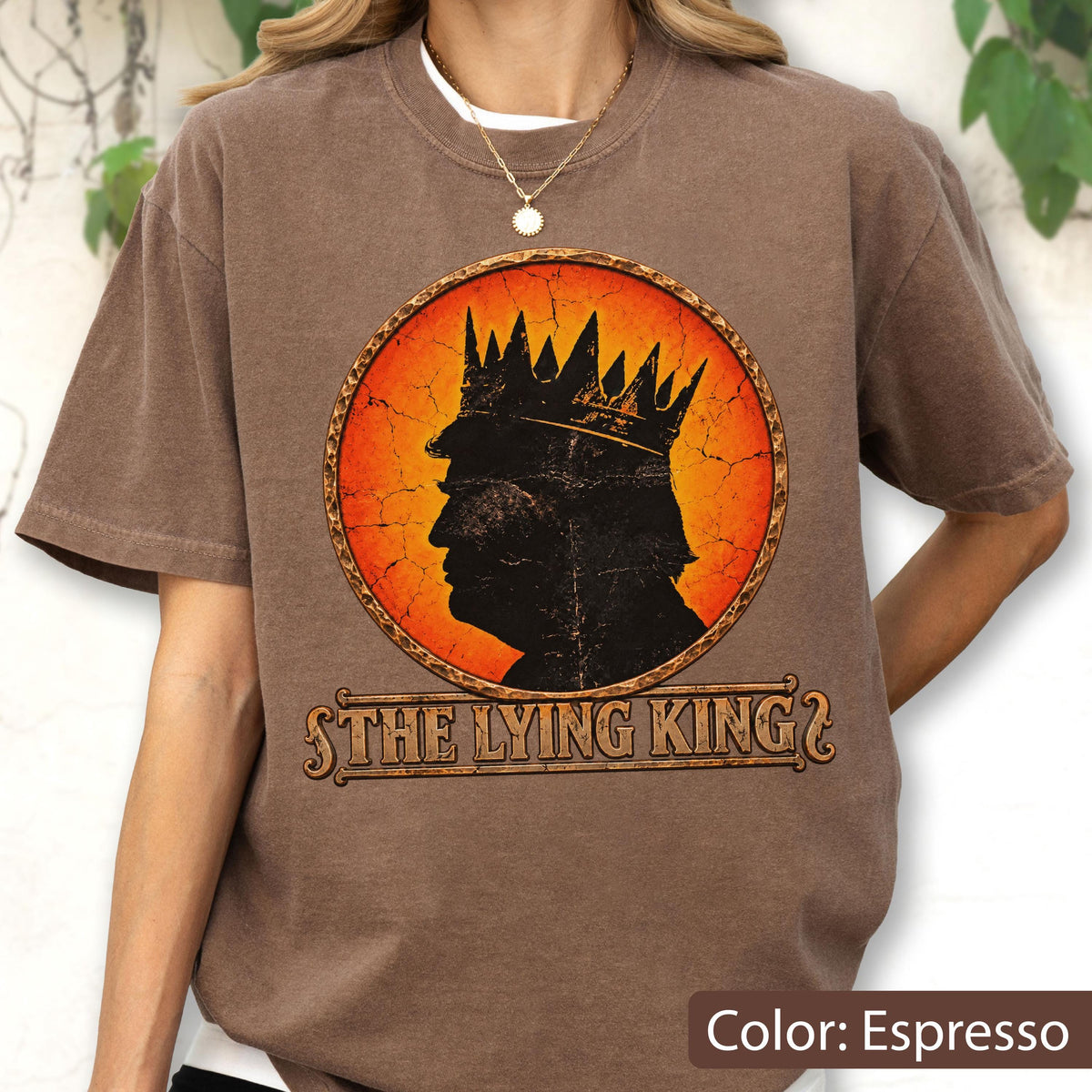 May include: An espresso brown t-shirt featuring a circular graphic with a cracked orange and red background. The graphic shows a black silhouette of a person wearing a crown. Below the circle, the text reads THE LYING KING in a stylized font.