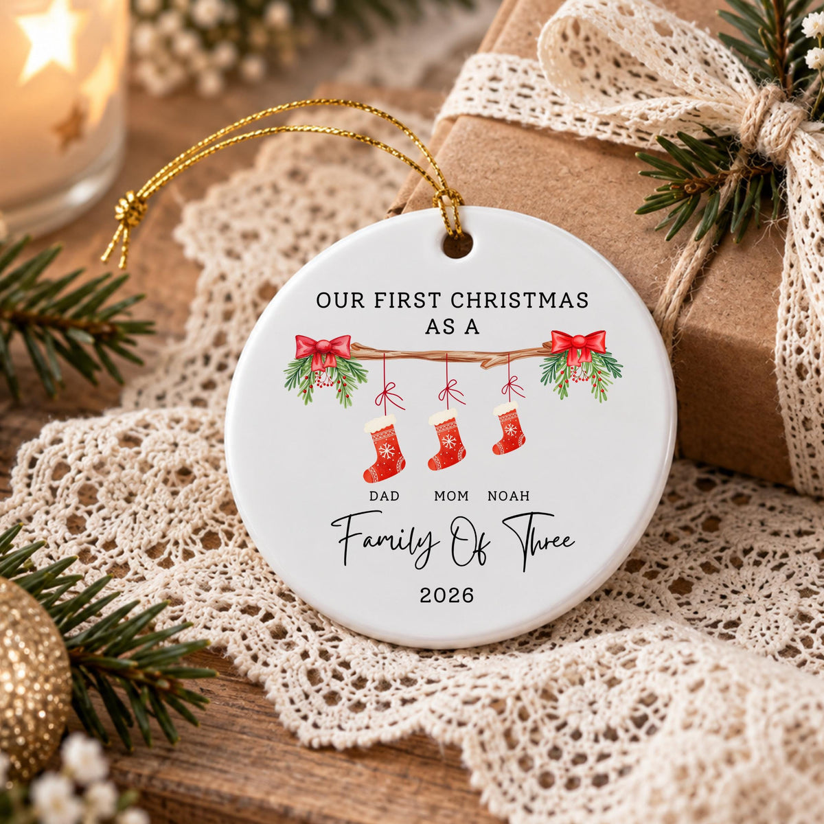 May include: A round white ceramic ornament featuring the text OUR FIRST CHRISTMAS AS A, followed by an illustration of a branch with three red stockings labeled DAD, MOM, and NOAH, and the text Family Of Three 2026, suspended by a gold string.