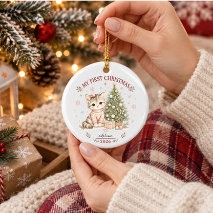 May include: A round white ceramic Christmas ornament featuring a graphic of a kitten wearing a pink sweater next to a decorated Christmas tree. The text reads MY FIRST CHRISTMAS, adeline, and 2026. It includes a gold hanging cord.
