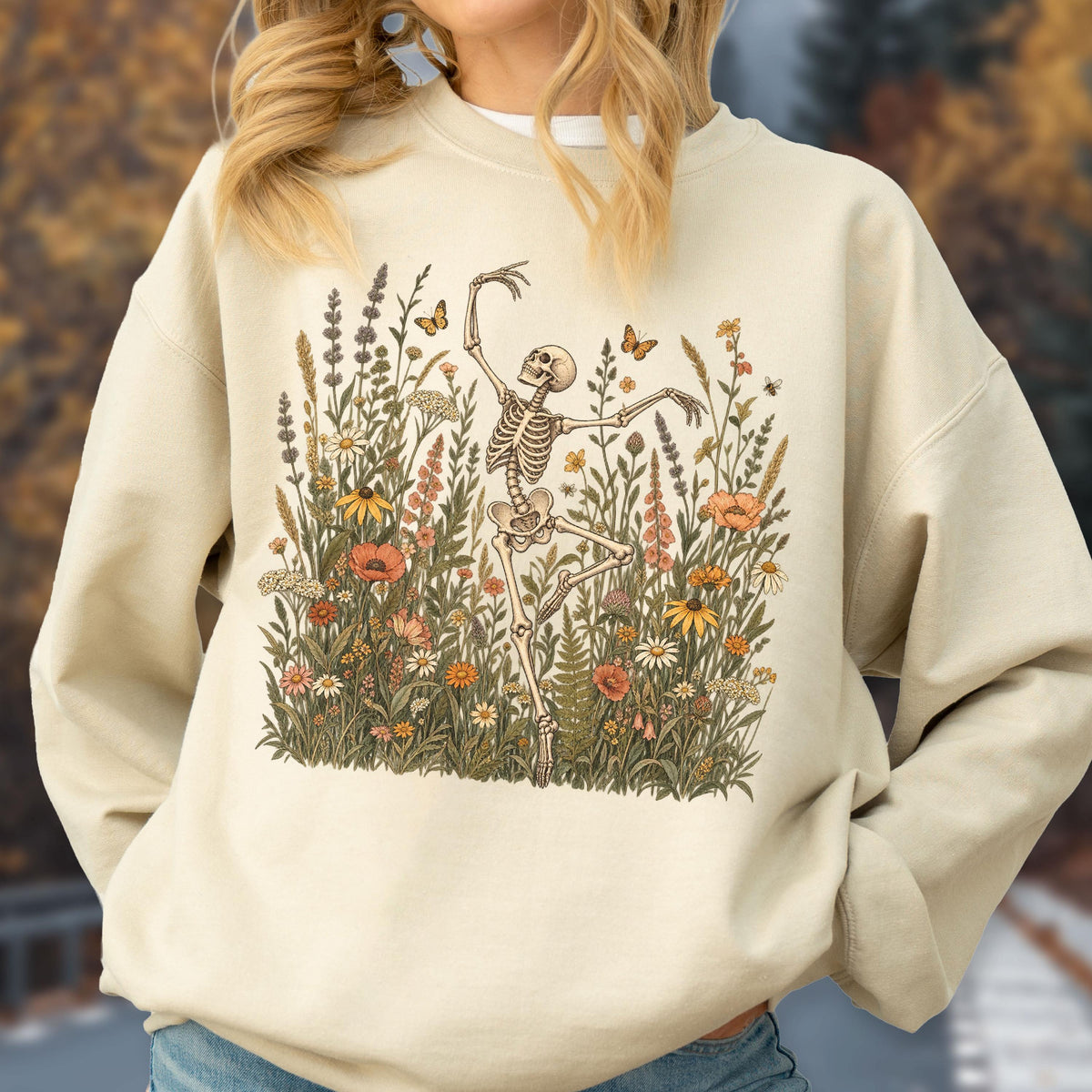 May include: A beige crewneck sweatshirt featuring a graphic print of a skeleton dancing amidst a field of colorful wildflowers, tall grasses, and small butterflies. The illustration uses muted tones of orange, yellow, green, and brown.