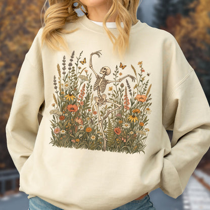 May include: A beige crewneck sweatshirt featuring a graphic print of a skeleton dancing amidst a field of colorful wildflowers, tall grasses, and small butterflies. The illustration uses muted tones of orange, yellow, green, and brown.