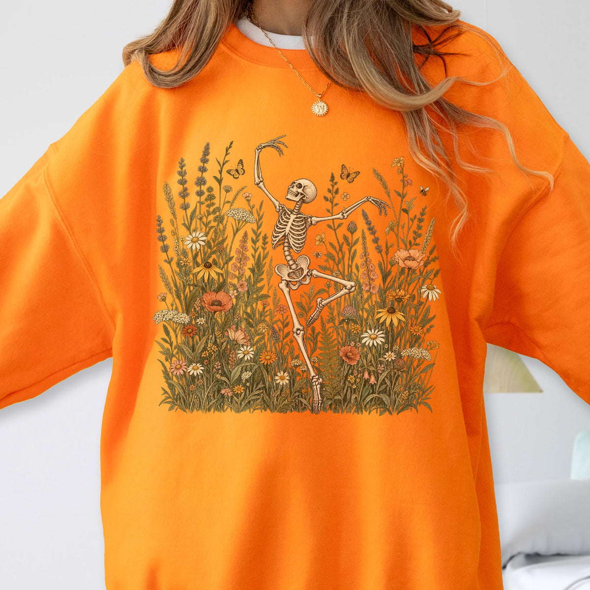 May include: A bright orange crewneck sweatshirt featuring a central graphic of a skeleton dancing amidst a field of colorful wildflowers, tall grasses, and small butterflies. The illustration uses earthy tones of brown, green, and muted red.