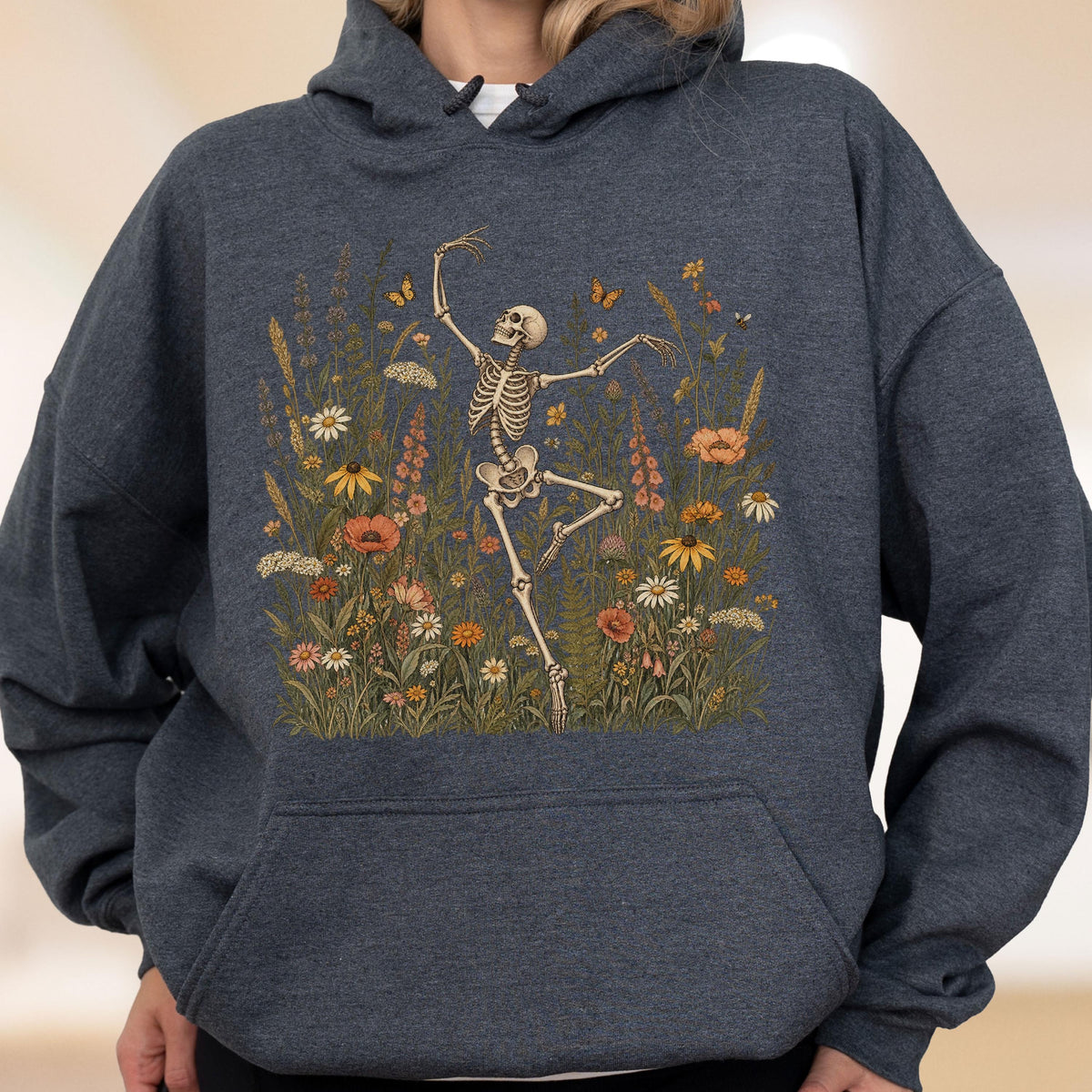 May include: A dark heather gray hooded sweatshirt featuring a graphic print of a human skeleton dancing amidst a field of colorful wildflowers, including daisies, poppies, and orange butterflies, with a front kangaroo pocket and long sleeves.