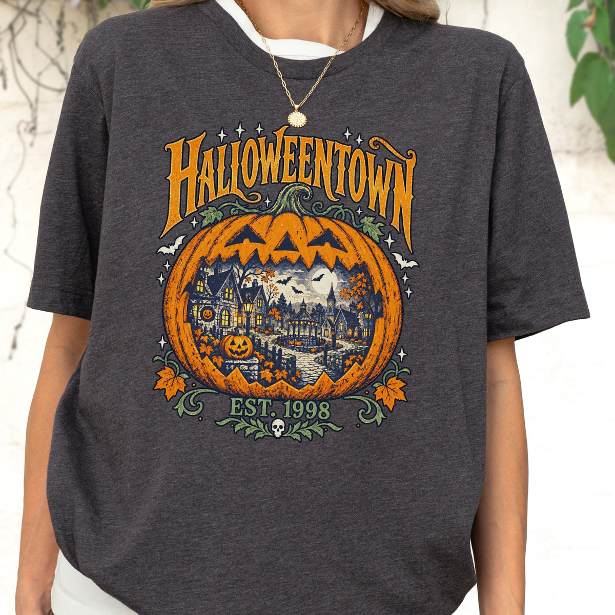 May include: A dark gray short-sleeve t-shirt featuring a large orange pumpkin graphic with a detailed scene of a house, trees, and bats inside. The text reads HALLOWEENTOWN and EST. 1998, accompanied by small stars, leaves, and a skull icon at the bottom.