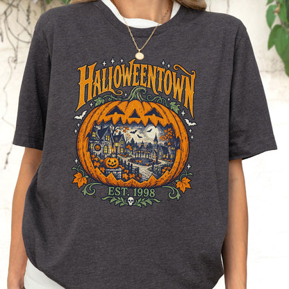 May include: A dark gray short-sleeve t-shirt featuring a large orange pumpkin graphic with a detailed scene of a house, trees, and bats inside. The text reads HALLOWEENTOWN and EST. 1998, accompanied by small stars, leaves, and a skull icon at the bottom.