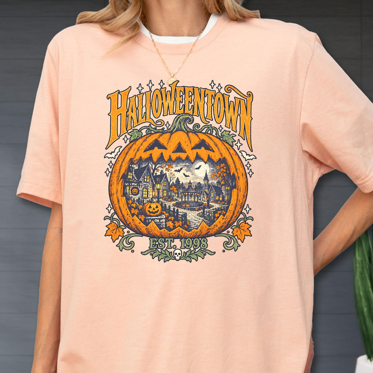May include: A peach-colored t-shirt featuring a graphic of a large orange jack-o'-lantern. Inside the pumpkin is a detailed scene of a spooky town with houses, trees, and a full moon. The text reads HALLOWEENTOWN and EST. 1998, with small bats and stars.