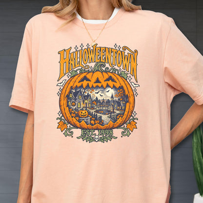 May include: A peach-colored t-shirt featuring a graphic of a large orange jack-o'-lantern. Inside the pumpkin is a detailed scene of a spooky town with houses, trees, and a full moon. The text reads HALLOWEENTOWN and EST. 1998, with small bats and stars.