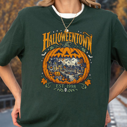 May include: A dark green t-shirt featuring a large orange jack-o'-lantern graphic with a detailed town scene inside. The text reads HALLOWEENTOWN and EST. 1998, accompanied by small bats, stars, and autumn leaves. The design is centered on the chest.
