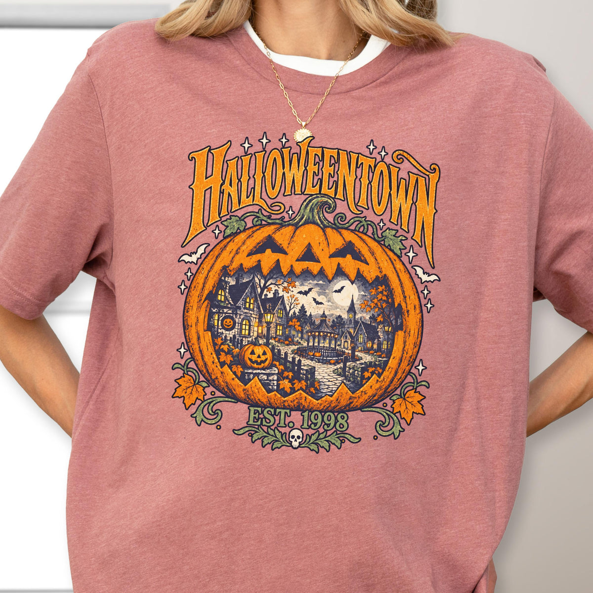 May include: A dusty rose short-sleeve t-shirt featuring a large graphic of a carved pumpkin with a spooky town scene inside. The text reads HALLOWEENTOWN and EST. 1998. The design includes orange, yellow, and dark blue colors with bats and leaves.