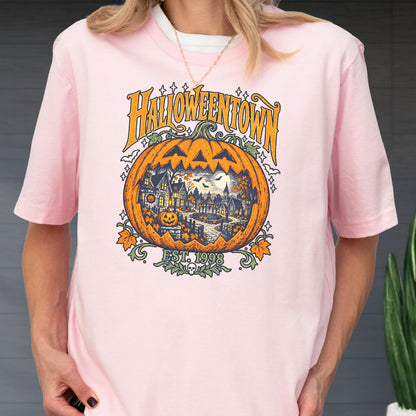 May include: A light pink short-sleeve t-shirt featuring a large graphic of a carved pumpkin with a spooky village scene inside. The text reads HALLOWEENTOWN and EST. 1998, accompanied by illustrations of bats, stars, and autumn leaves in orange and black.