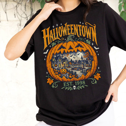 May include: A black t-shirt featuring a large orange pumpkin graphic with a detailed scene of a town inside it. The text HALLOWEENTOWN is printed in yellow above the pumpkin, with EST. 1998 and a small skull icon printed in green below it.