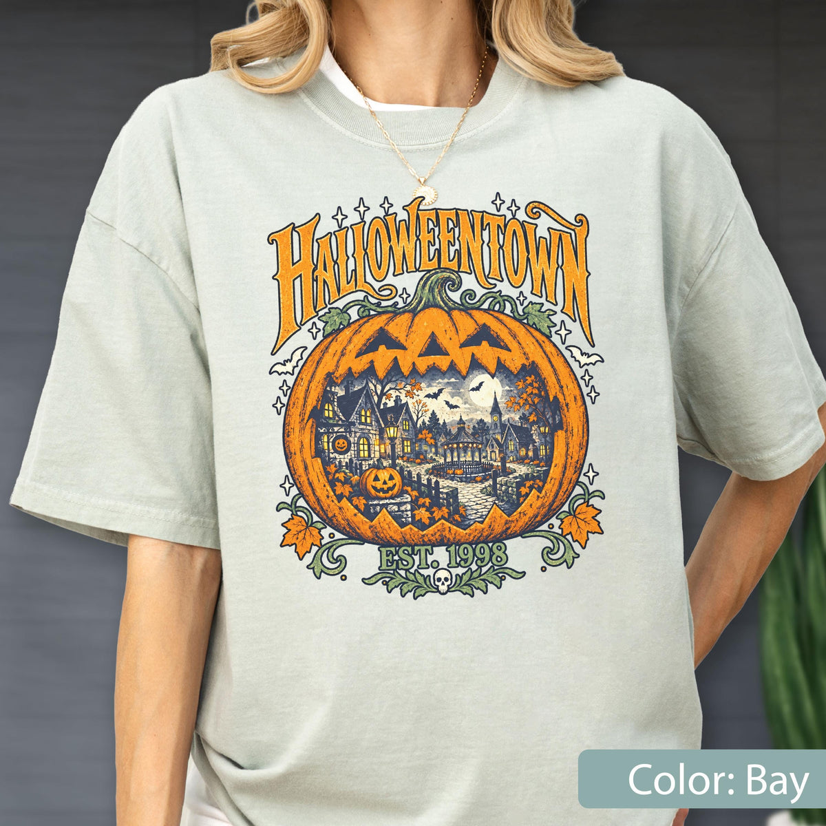 May include: A light sage green short-sleeve t-shirt featuring a large graphic print of a carved pumpkin with a town scene inside. The text HALLOWEENTOWN is arched above the pumpkin, with EST. 1998 and a small skull icon printed below it.