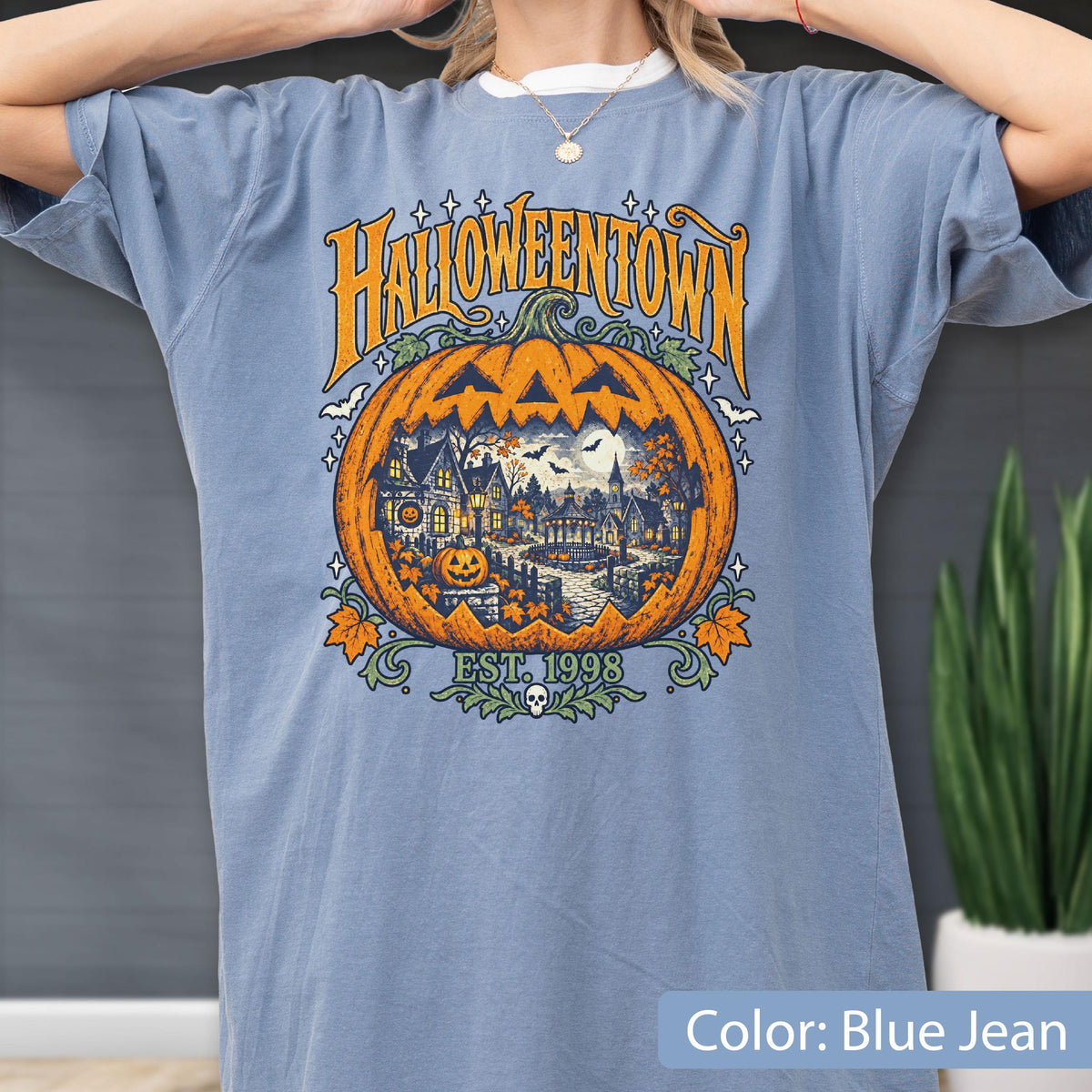May include: A blue jean colored t-shirt featuring a large graphic of a carved pumpkin containing a detailed scene of a town with houses, trees, and bats. The text HALLOWEENTOWN is printed in orange above the pumpkin, with EST. 1998 below it.