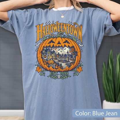 May include: A blue jean colored t-shirt featuring a large graphic of a carved pumpkin containing a detailed scene of a town with houses, trees, and bats. The text HALLOWEENTOWN is printed in orange above the pumpkin, with EST. 1998 below it.