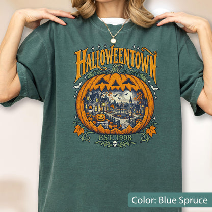 May include: A blue spruce t-shirt featuring a large orange jack-o'-lantern graphic with a detailed town scene inside. The text reads HALLOWEENTOWN and EST. 1998, accompanied by small bats, stars, and a skull icon at the bottom of the design.