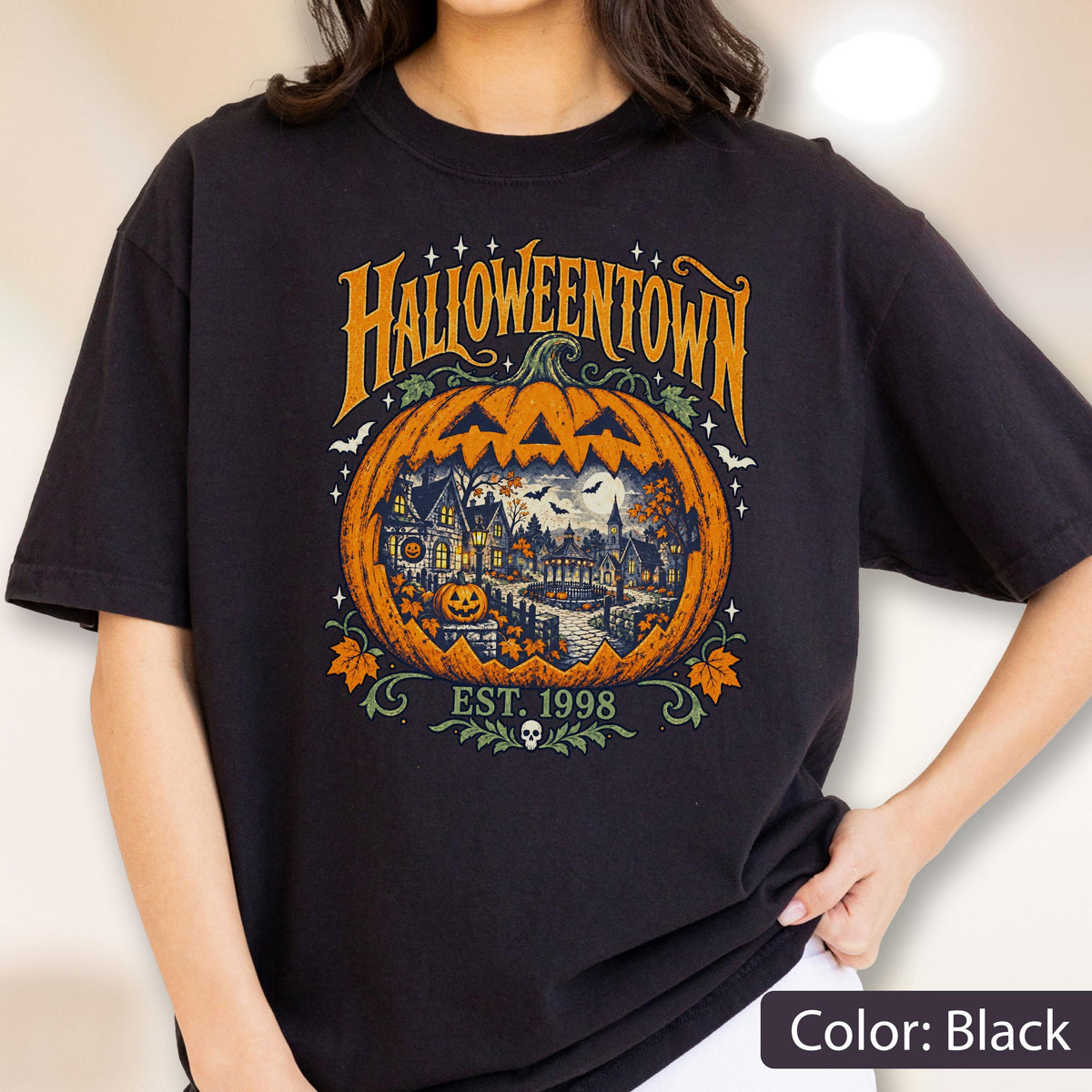May include: A black t-shirt featuring a large orange pumpkin graphic with the text HALLOWEENTOWN and EST. 1998. The pumpkin contains a detailed illustration of a spooky village scene with houses, trees, bats, and a full moon, accented by small leaves.