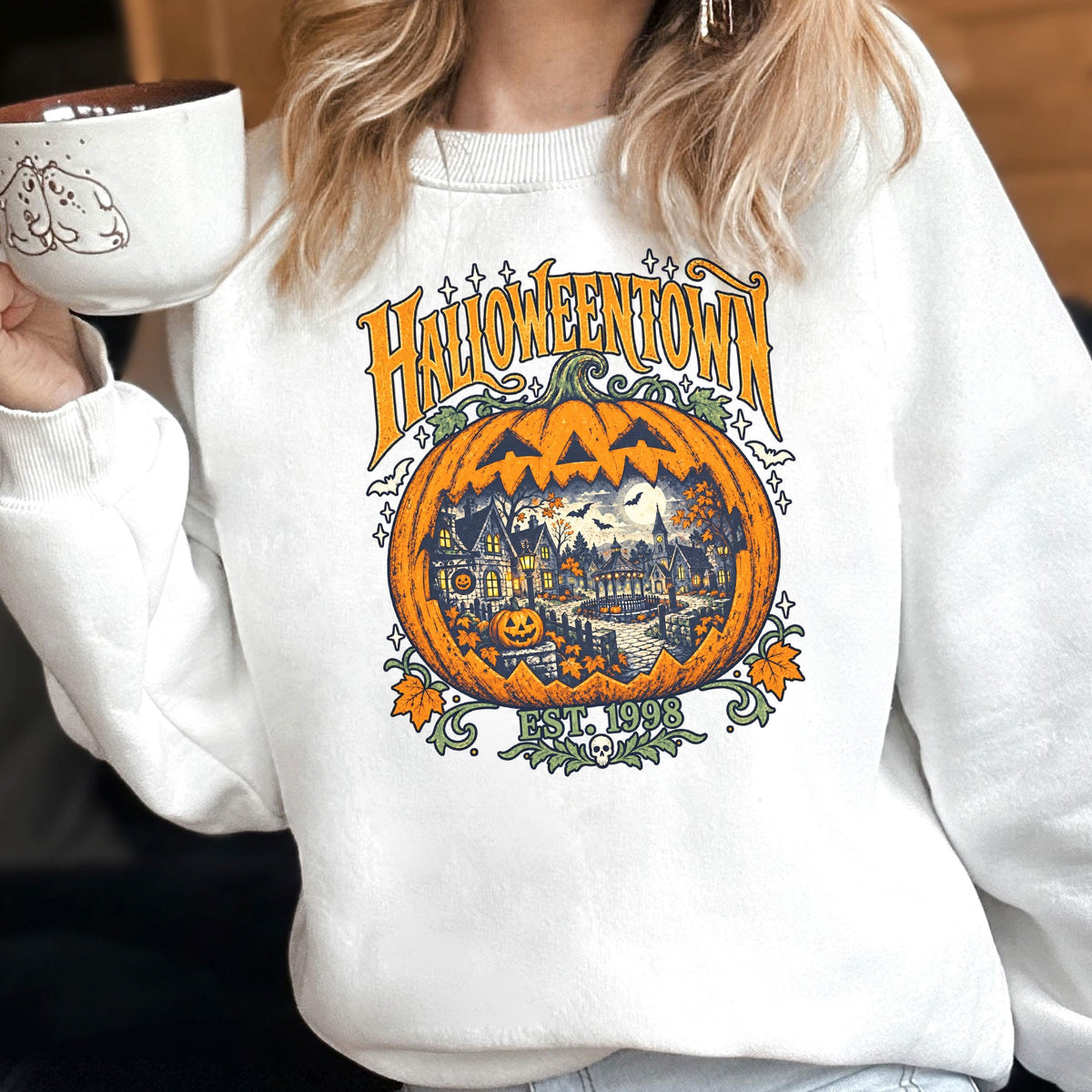 May include: A white crewneck sweatshirt featuring a graphic print of a large orange jack-o'-lantern containing a detailed scene of a spooky town. The text HALLOWEENTOWN is printed in orange above the pumpkin, with EST. 1998 and a small skull below.