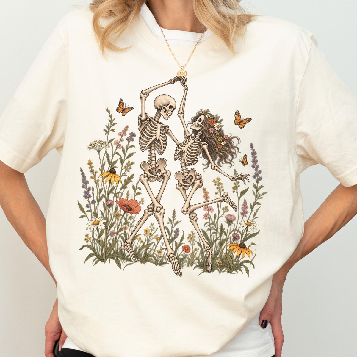 May include: A cream-colored t-shirt featuring a graphic print of two skeletons dancing in a field of colorful wildflowers and butterflies. The skeletons are positioned centrally, surrounded by various botanical elements including daisies, poppies, and tall grasses, with orange butterflies fluttering nearby.