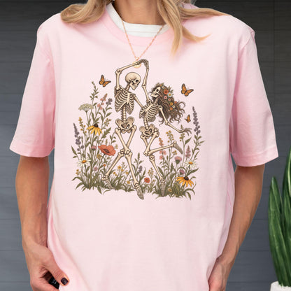 May include: A light pink short-sleeve t-shirt featuring a graphic print of two skeletons dancing in a field of wildflowers. The illustration includes various colorful flowers, green foliage, and several orange butterflies surrounding the skeletons.