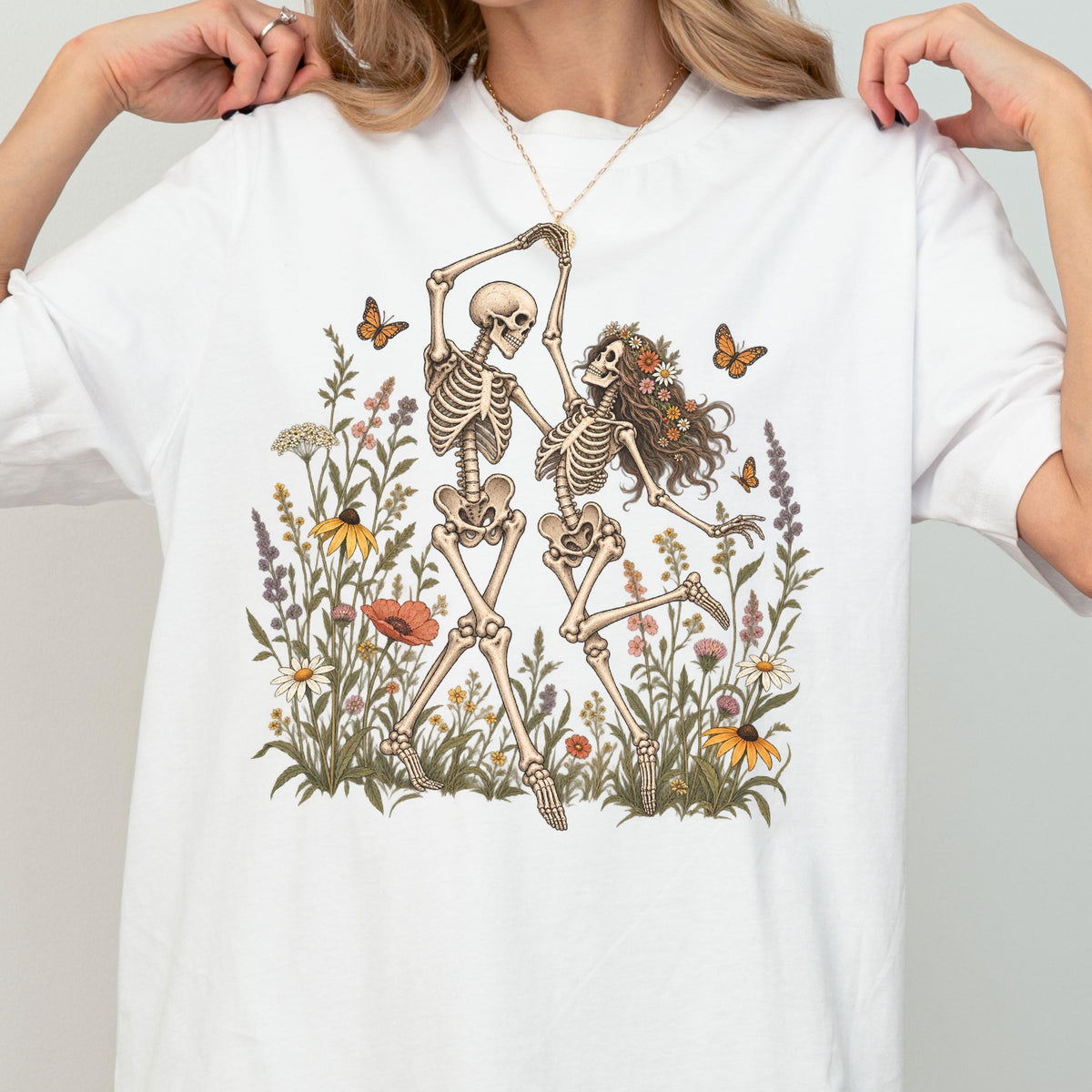 May include: A white t-shirt featuring a graphic illustration of two skeletons dancing amidst a field of colorful wildflowers and butterflies. One skeleton has long hair adorned with a floral crown, while the other is bare-headed.