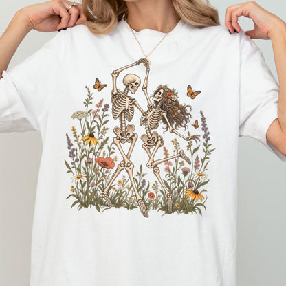 May include: A white t-shirt featuring a graphic illustration of two skeletons dancing amidst a field of colorful wildflowers and butterflies. One skeleton has long hair adorned with a floral crown, while the other is bare-headed.