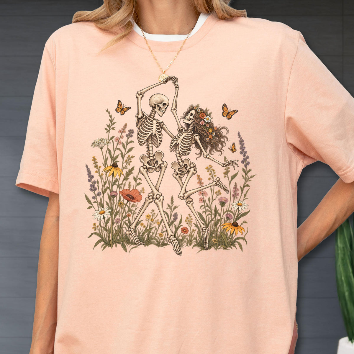 May include: A peach-colored t-shirt featuring a graphic print of two skeletons dancing amidst a field of colorful wildflowers and butterflies. The skeletons are depicted in a light brown ink, while the surrounding floral elements include various blossoms.