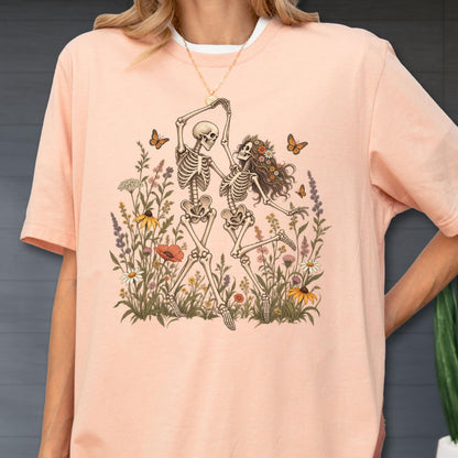 May include: A peach-colored t-shirt featuring a graphic print of two skeletons dancing amidst a field of colorful wildflowers and butterflies. The skeletons are depicted in a light brown ink, while the surrounding floral elements include various blossoms.