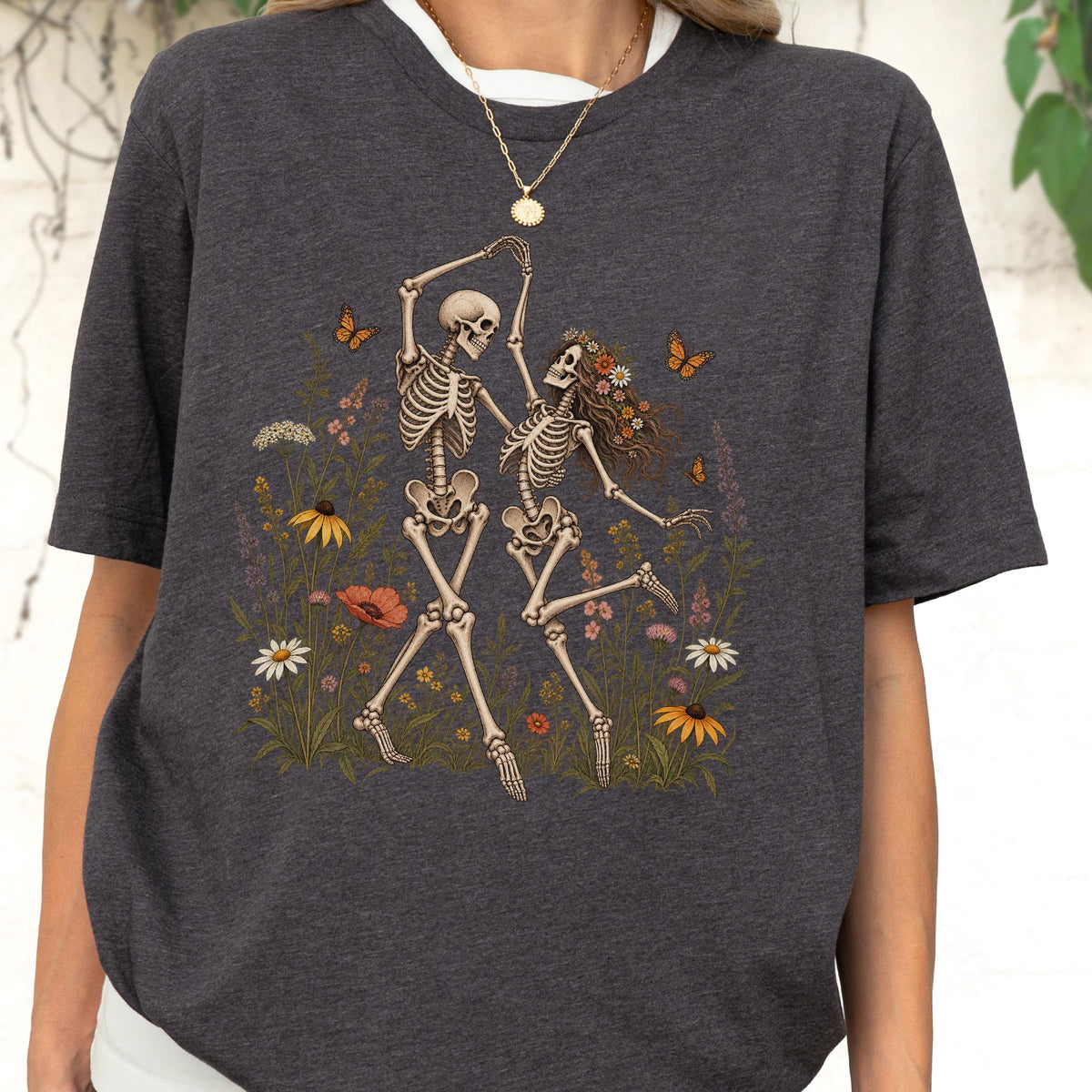 May include: A dark charcoal gray short-sleeve t-shirt featuring a graphic print of two skeletons dancing in a field of colorful wildflowers. The skeletons are surrounded by various blossoms, green foliage, and small orange butterflies in flight.