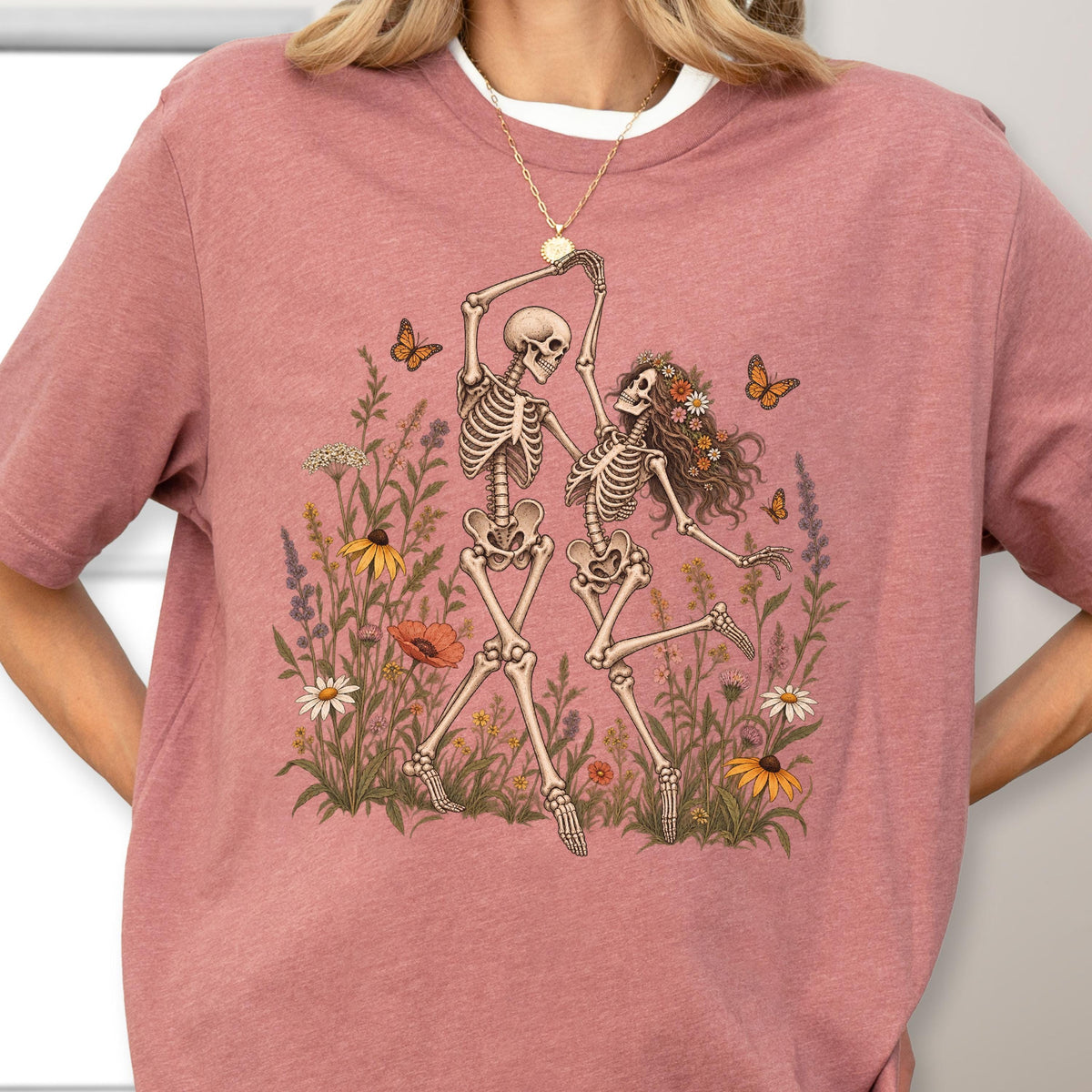 May include: A dusty rose short-sleeved t-shirt featuring a graphic print of two skeletons dancing in a field of wildflowers. The skeletons are surrounded by colorful flowers and orange butterflies. The shirt has a crew neckline and a relaxed fit.