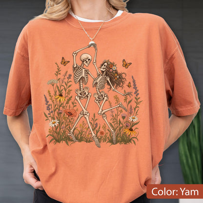 May include: A yam-colored t-shirt featuring a graphic print of two skeletons dancing amidst a field of colorful wildflowers and butterflies. The skeletons are depicted in a light beige tone, surrounded by various green stems and vibrant floral accents.