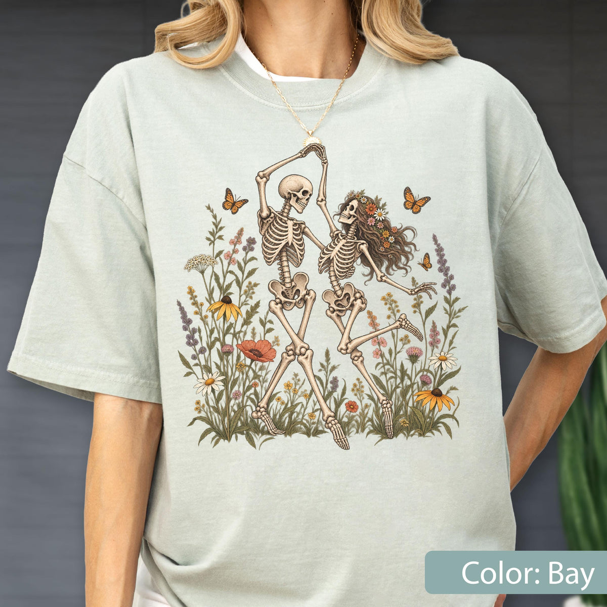May include: A light sage green t-shirt featuring a graphic illustration of two skeletons dancing in a field of colorful wildflowers and butterflies. The skeletons are depicted in a joyful pose, surrounded by various botanical elements and insects.