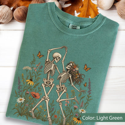 May include: A light green t-shirt featuring a graphic of two skeletons dancing among colorful wildflowers and butterflies. The skeletons are positioned in the center, surrounded by various floral elements. The shirt has a Comfort Colors brand label.
