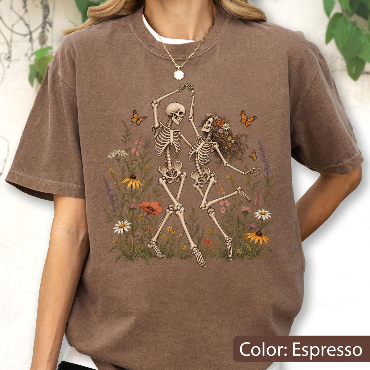 May include: An espresso-colored t-shirt featuring a graphic print of two skeletons dancing amidst a field of colorful wildflowers and orange butterflies. The design includes various floral elements in shades of yellow, red, and white.